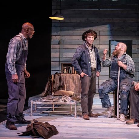 Four actors are gathered onstage discussing an important topic. The two actors on the left are stood, the two actors on the right are sat as the conversation takes place.