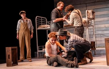 Five actors are shown onstage. The two upstage are crouched and appear to be having a serious discussion. Two actors are stood behind them, one has a cut hand and the other holds a rag attempting to stop the bleeding. The final actor is stood to the left of the screen and is onlooking.