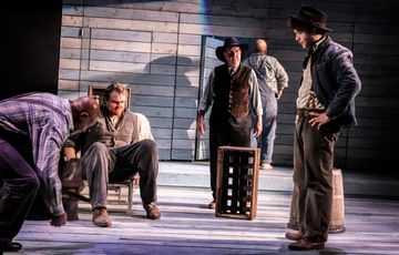 The actors are spread around the stage. The focus is one actor sat in a chair. He looks angry, confused and serious