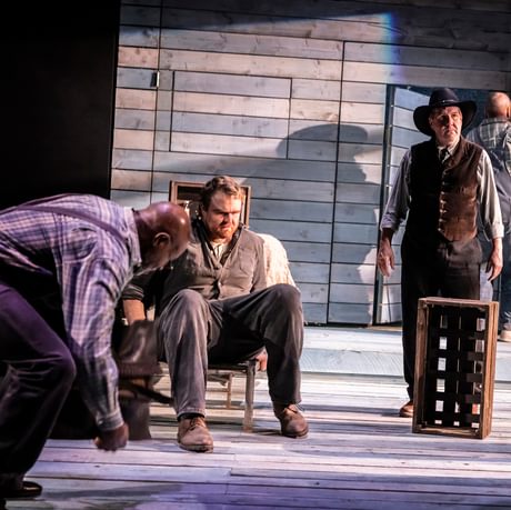 The actors are spread around the stage. The focus is one actor sat in a chair. He looks angry, confused and serious