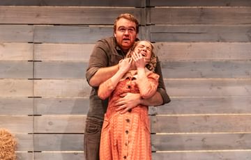 Two actors are shown on stage. One actor has a hold of the other from behind and appears to be restraining her out of fear.