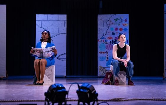 Two actors on stage sat on blocks. They look ahead into the distance