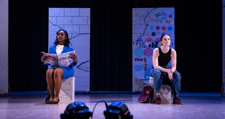 Two actors on stage sat on blocks. They look ahead into the distance