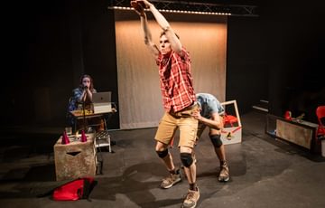 Two actors are creating the shape of a dinosaur. On stands upright with his hands over his head, whilst the other is bent over behind him.