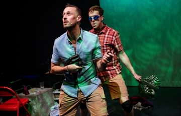 The actors are on stage stood one in front of the other. The actor behind wears goggles and is holding tree leaves in his hands