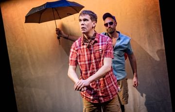 The actors are on stage. The actor downstage looks worried, whilst the actor upstage is wearing a cap, sunglasses and holding an opened umbrella.