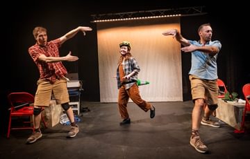 Three actors are on stage. The two actors either side of the stage hold their hands inwards, whilst the actor in the centre wears a bike helmet on his head, and a green traffic cone on his back.