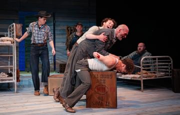 Actors are spread across the stage. A fight appears to have broken out between the actors upstage. One actor pins another actor down, whilst a third one attempts to pull him off.