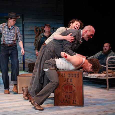 Actors are spread across the stage. A fight appears to have broken out between the actors upstage. One actor pins another actor down, whilst a third one attempts to pull him off.