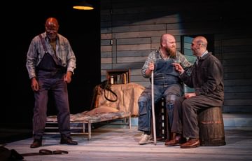 Ben Wilson, playing Candy, and Luke Richards, playing Lennie sit laughing together as Jeff Atkinson, playing Crooks, stands to their left smiling.
