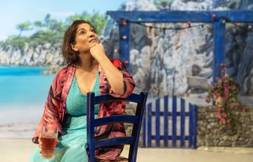 The actress is sat in front of a Greece setting. She is sat on a chair holding a drink and looking up into the distance.
