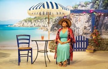 The actress is in front of a Greece setting, she is wearing beach attire and is sat at a table with a drink