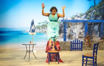 The actress is in front of a Greece setting, she stands on top of a chair and is about to jump off of it.