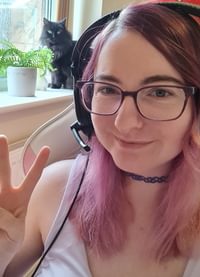 White female sat down, smiling, looking directly to camera, displaying the peace sign with her fingers. She has long light pink hair, wearing glasses and has headphones on. She is sat next to an open window which has some potted plants and a cat sat on the window ledge.