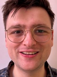 Head and shoulders image of a white male looking directly at camera smiling. He has brown hair, glasses and a patterned shirt