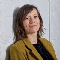 Female standing with back against the wall looking directly at camera and smiling. In a black blouse and mustard blazer