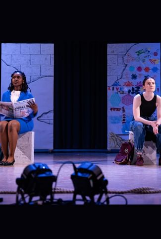 Two actors on stage sat on blocks. They look ahead into the distance