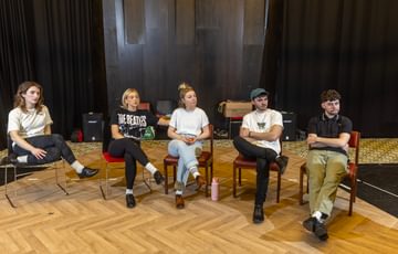 Five actors are shown sat on chairs with their legs crossed as they look off to the right of the screen