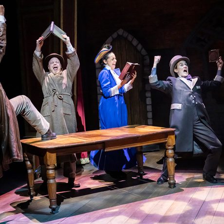 4 actors in Victorian dress holding arms aloft and celebating