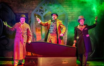 3 actors holding arms aloft over a an open coffin