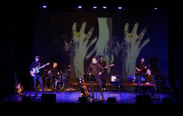 A band consisting of 6 people stand/sit on a lit stage. All playing different instruments such as: Guitar, Keyboard, Drums, Bass and one person looks like they will sing. The back wall has some large hands projected on to it.