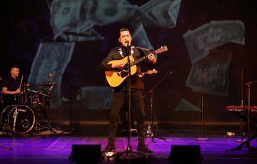 A person stands centre stage in a purple hue, they are playing the guitar and singing. The back wall of the stage has falling dollar bills projected onto it.