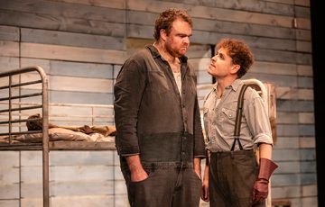 William Young, playing Lennie stands with his hands in his pockets as Stefan Race, playing Curley, angrily looks up to him.