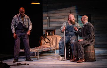 Ben Wilson, playing Candy, and Luke Richards, playing Lennie sit laughing together as Jeff Atkinson, playing Crooks, stands to their left smiling.