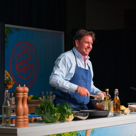 A man stood behind a kitchen surface cooking. He is wearing a pale blue shirt, a dark blue apron and is smiling.