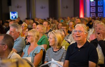 A crowd of people sitting, all looking the same direction. Some are laughing, some are smiling and one is yawning.