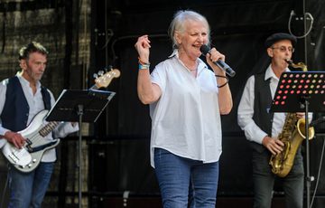 A lady, wearing a white blouse and blue wash jeans, is holding a microphone. To her left is a man playing a bass guitar, to her right, a man blowing into a saxaphone.