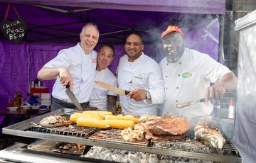 Four chefs stand behind a grill, an array of different foods is cooking on the grill. They are all smiling.