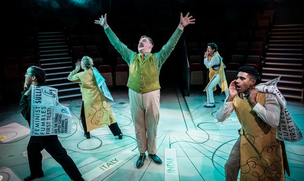 A man wearing a green waistcoat throws his hands in the air. He is surrounded by people carrying large newspapers.
