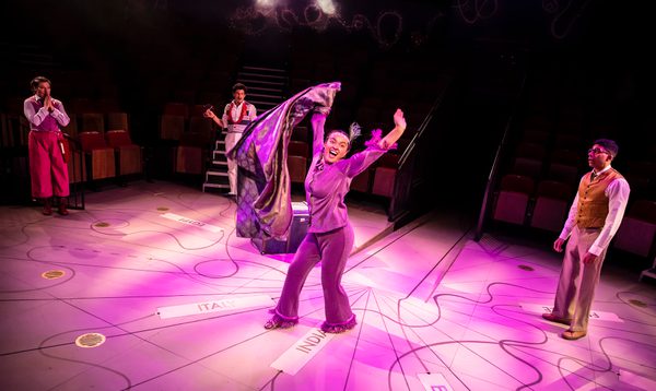 A woman wears a sparkly purple jumpsuit, with a purple feather in her hair - she looks extremely happy.