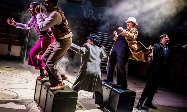 6 people, 3 of which are stood on chests. Two of them wear long trench coats and trilby's.