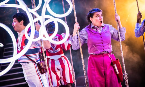 A woman in a purple waistcoat holding on to a swing. Neon tubes shine a white light to the left of the picture, a man's head with brown hair can be seen between the lights.