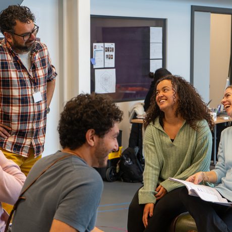 Group of 5 men and women sat in a rehearsal room. Smiling and chatting with some holding scripts.