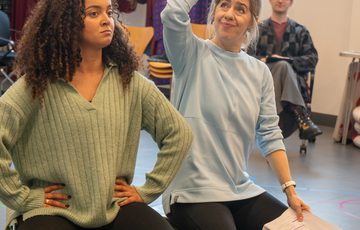 Two women sat on a luggage trunk. One with hands on hips another holding a script in one hand and other hand on her head.