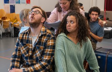 A man and a woman sat on a luggage trunk with bemused expressions. A woman stood behind them looking at a script.