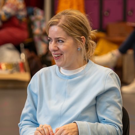 A woman sat down on a chair with a folder open on her lap, she is smiling and looking at something off camera.