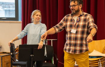 A woman stood leaning on two music stands and a man stood gesturing with both hands out in front of him.