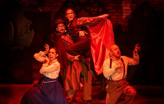 Image of 4 actors on stage with a red light, one of the actors is dressed as Dracula