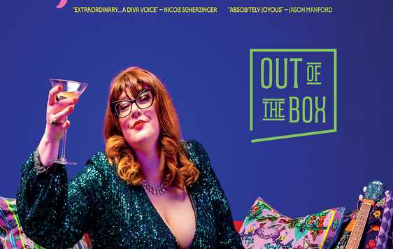 A woman with ginger hair, wearing glasses and a green sequin dress sat on a sofa with a martinin glass held up in the air