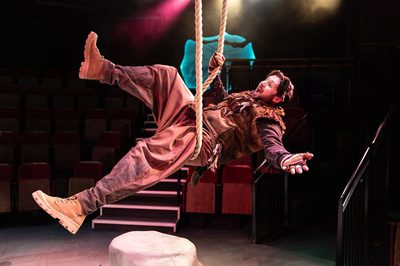 A man in a brown costume lying on a rope swing with pink and yellow lights in the background.