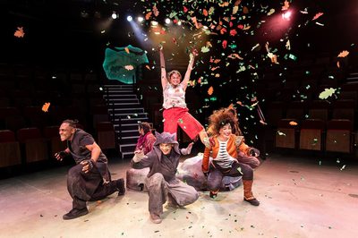 A woman standing on a rock with her hands up. Five people squatting around the rock with orange and green confetti flying around them and yellow, white, and red lights in the background.