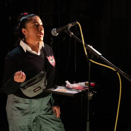 A young brown woman hair tied back with a braid, wearing a black top and green trousers and a crossbody bag talking into a mic.