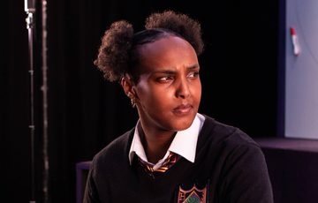 A young black women sat down. She has hair tied back, wearing white shirt and black school jumper