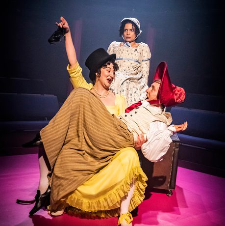 3 actors on stage, man wearing a dress sitting on a womans lap.