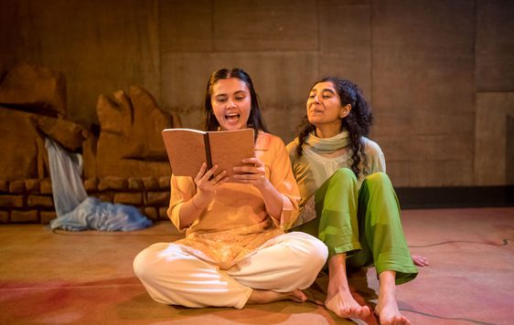 Two girls sat on the floor, one absorbed in a book while the other is listening attentively.