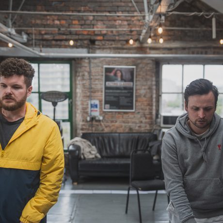 Two men looking sad. Man 1 has short curly hair and a beard he is wearing a yellow and man 2 has straight short hair and is wearingblue jacket and the other is wearing a grey jumper